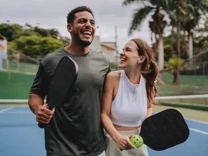 Two players laughing together on an outdoor pickleball court, paddles in hand.
